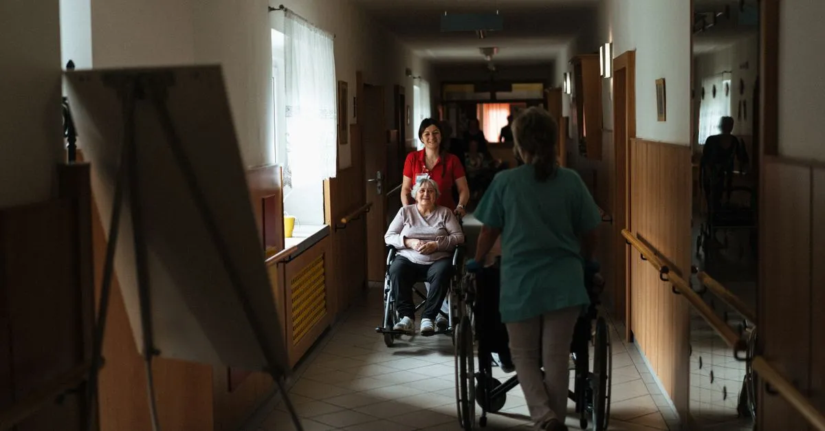 Pflegerin begleitet ältere Frau im Rollstuhl durch einen Korridor eines Pflegezentrums