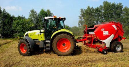 Grüner Traktor mit Heuballen-Presse auf einem Feld mit Bäumen im Hintergrund