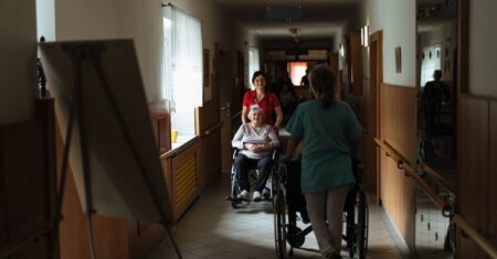 Pflegerin begleitet ältere Frau im Rollstuhl durch einen Korridor eines Pflegezentrums